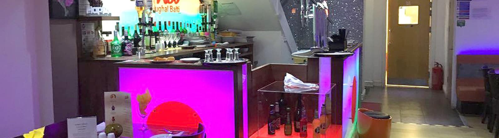 The bar area, lit in pink and purple, with bottles and glassware on the counter.
